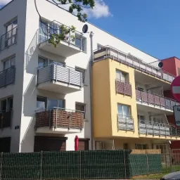 Nowoczesny, wielobarwny budynek mieszkalny z balkonami, widoczny z poziomu ulicy, z zielonym ogrodzeniem i znakiem drogowym na pierwszym planie.