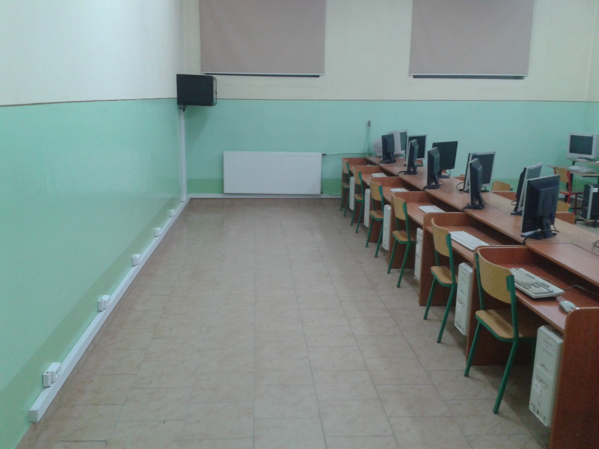 Sala komputerowa z rzędem biurek, komputerami stacjonarnymi i krzesłami, widoczne listwy przypodłogowe z gniazdkami elektrycznymi wzdłuż ściany.