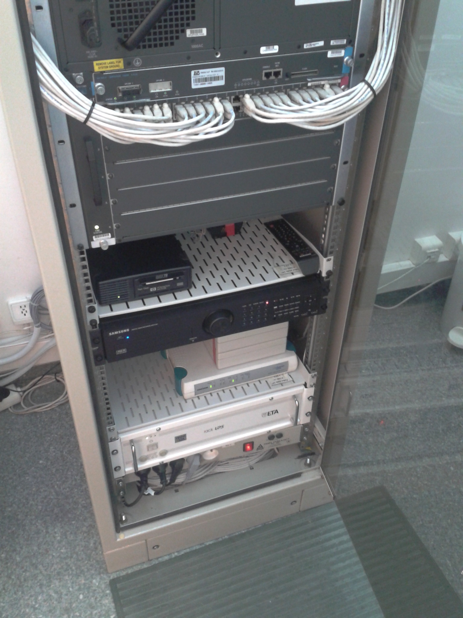 Otwarta szafa rack z zamontowanymi urządzeniami sieciowymi i serwerowymi, widoczne kable, zasilacze UPS, przełączniki, routery i inne urządzenia, na podłodze szary dywan.