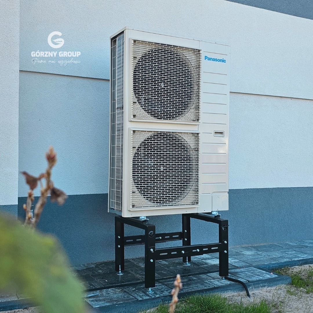 Biała pompa ciepła Panasonic na czarnym metalowym stelażu, zainstalowana przy jasnej ścianie budynku z widocznym logo firmy Górny Group w tle.