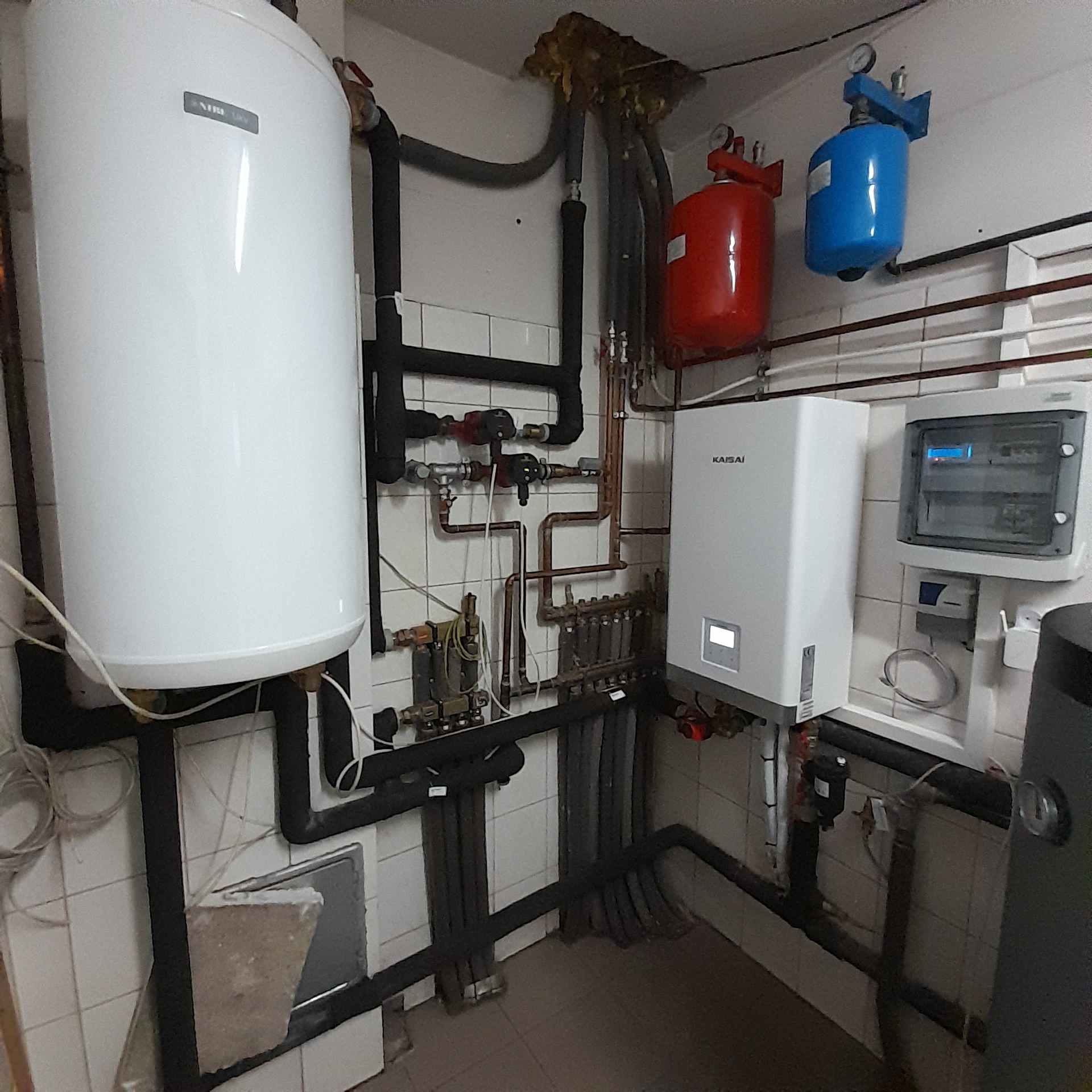 Kotłownia z białym zasobnikiem ciepłej wody, kotłem gazowym Kaisai, czerwonym i niebieskim naczyniem wzbiorczym, oraz rozbudowaną instalacją hydrauliczną z rur miedzianych i czarnych izolowanych.