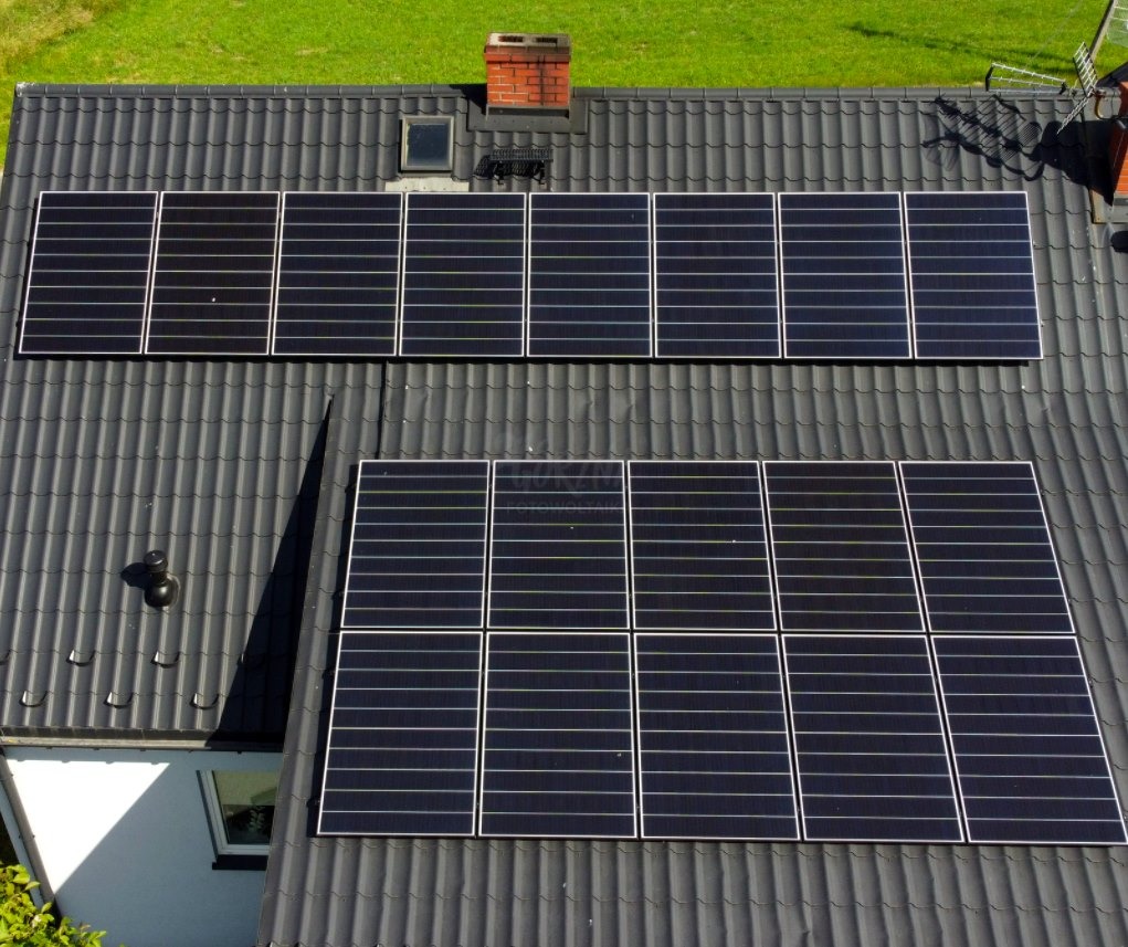 Widok z góry na panele słoneczne zamontowane na dachu domu pokrytym szarą dachówką, z kominem w tle i fragmentem zielonego trawnika.