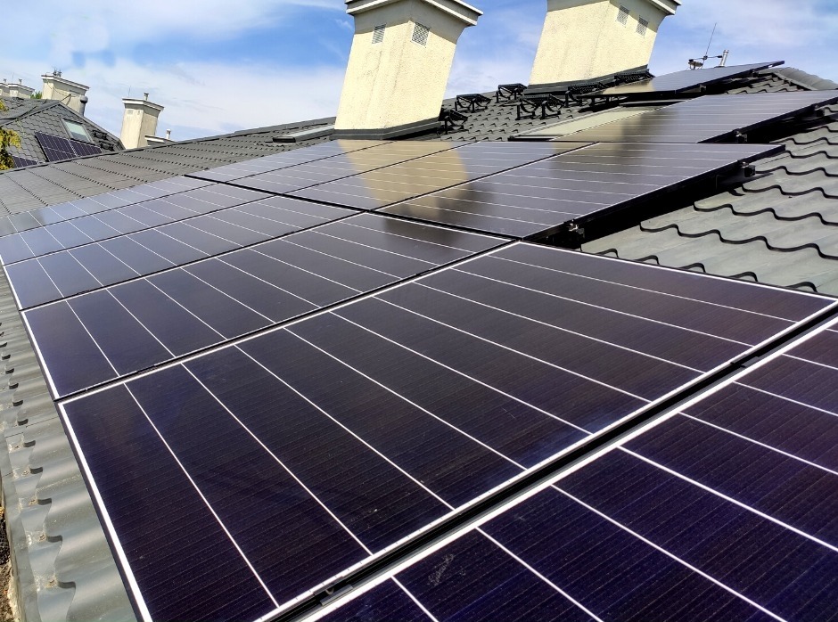 Panele słoneczne na ciemnym, metalowym dachu domu z kominami, widoczne z bliska w słoneczny dzień.