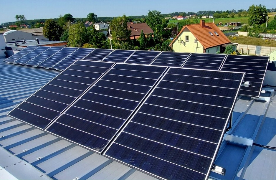 Rząd paneli słonecznych na metalowym dachu z widokiem na okoliczne domy i drzewa w słoneczny dzień.