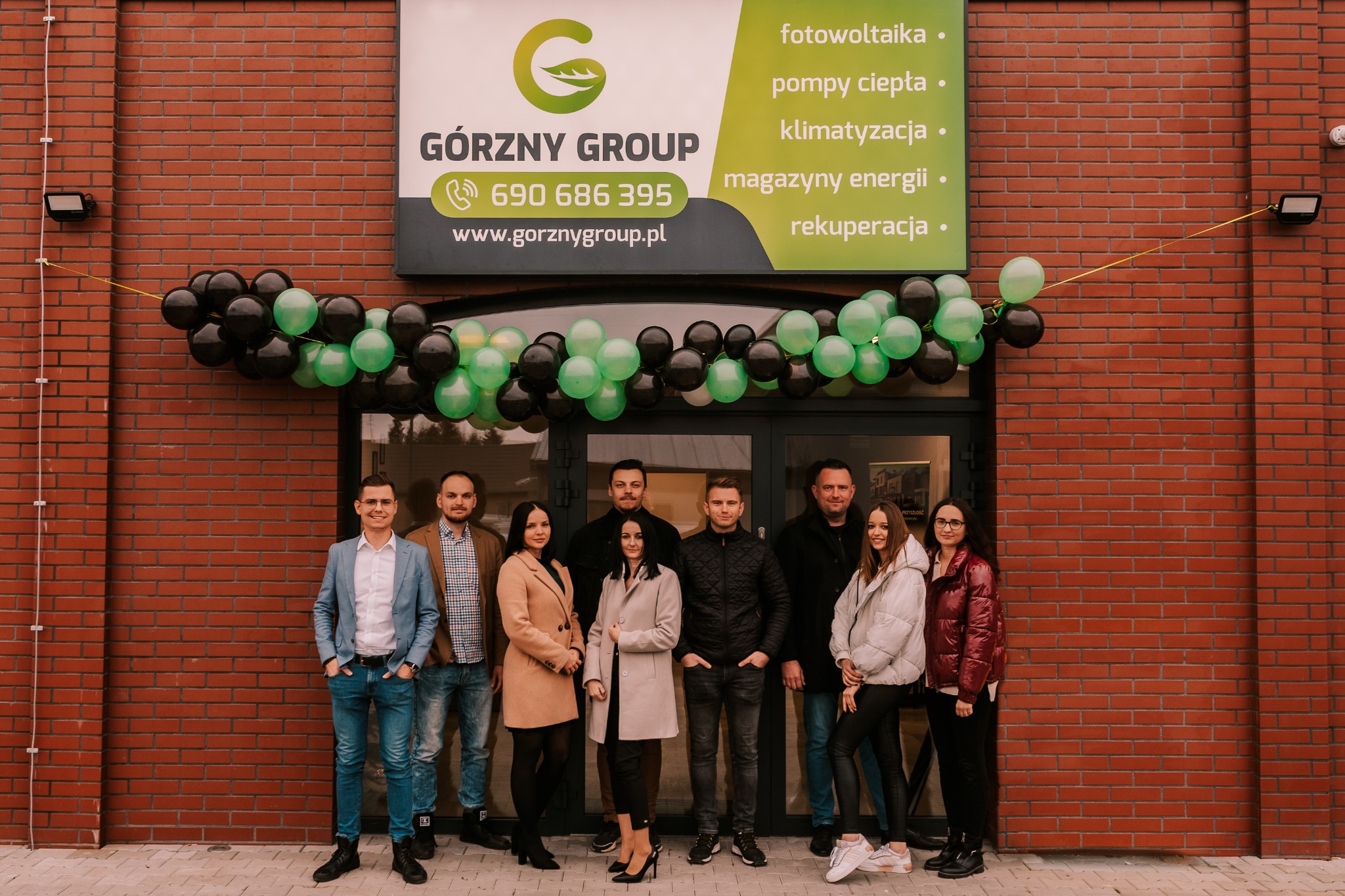 Grupowe zdjęcie zespołu Górzny Group przed budynkiem z szyldem oferowanych usług: fotowoltaika, pompy ciepła, klimatyzacja, magazyny energii i rekuperacja, udekorowanym czarno-zielonymi balonami.