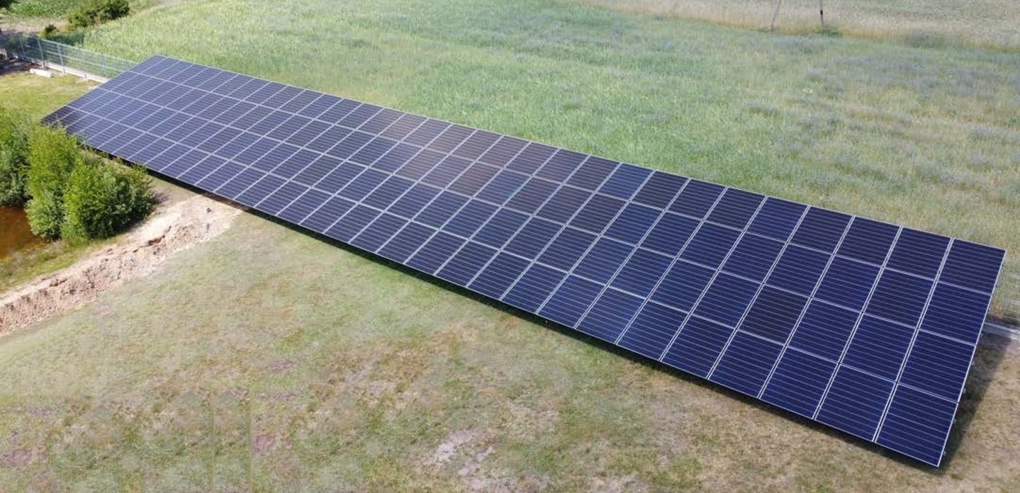 Rozległa instalacja paneli słonecznych na trawiastym terenie, widok z góry, obok widoczny niewielki zbiornik wodny i ogrodzenie.