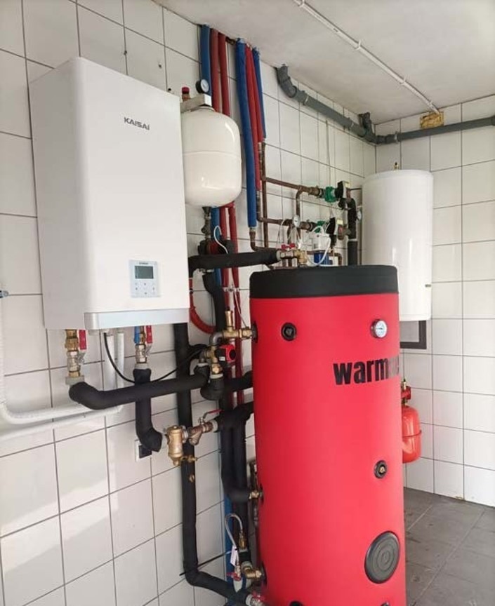 Instalacja grzewcza w kotłowni: biały moduł Kaisai, czerwony zasobnik ciepłej wody Warm, rury w izolacji, białe płytki na ścianach.