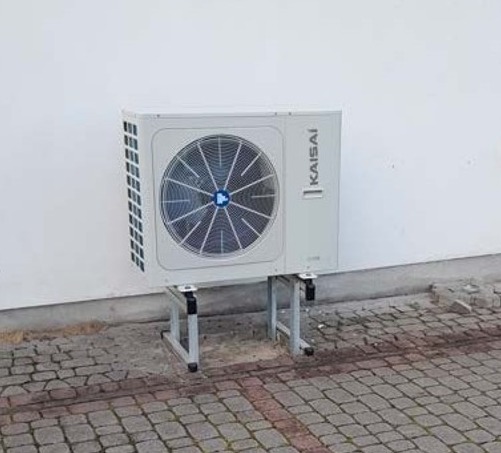 Biała jednostka zewnętrzna klimatyzacji marki Kaisai zamontowana na metalowym stelażu przy białej ścianie budynku, widok z dołu na tle brukowanej nawierzchni.