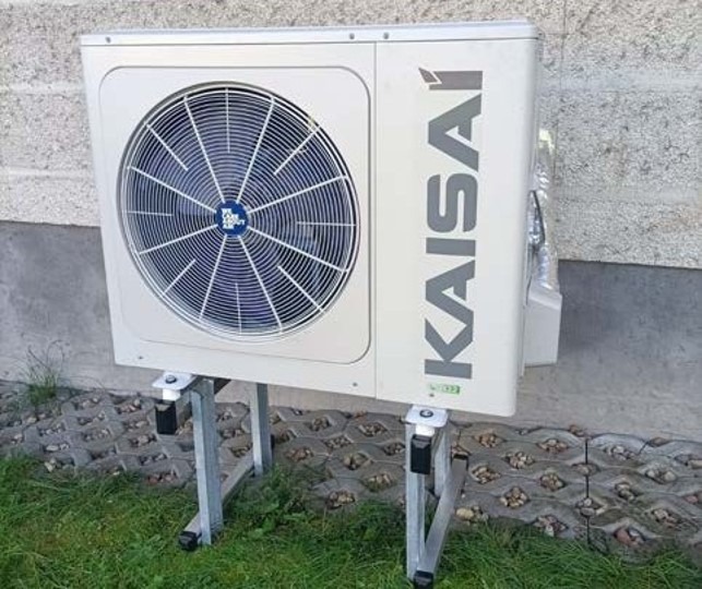 Zewnętrzna jednostka pompy ciepła marki Kaisai zamontowana na metalowym stelażu przy ścianie budynku z kostki betonowej, otoczona trawnikiem.