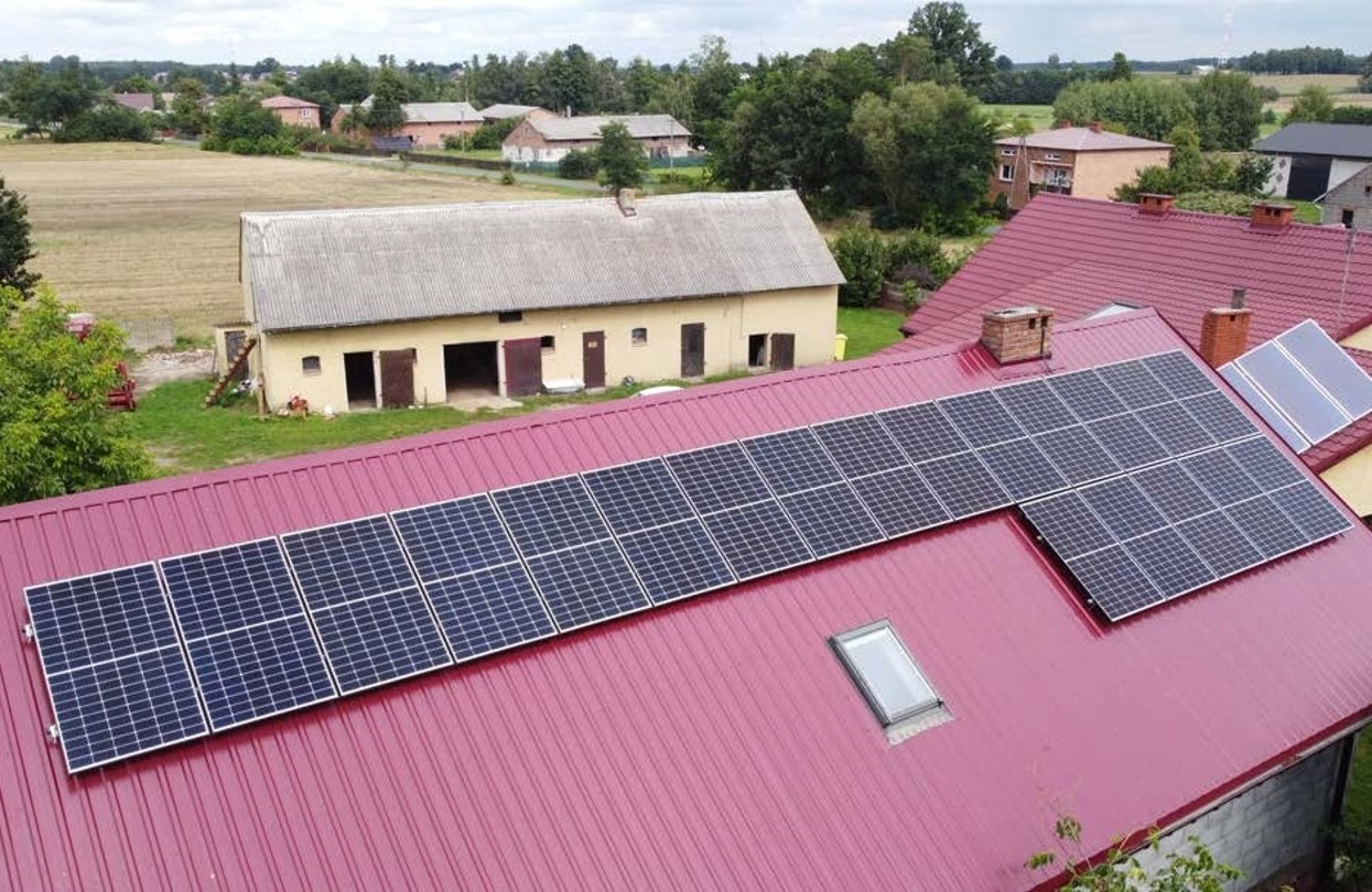 Instalacja paneli słonecznych na dachu domu z blachy w kolorze czerwonym, widok z góry na tle zabudowań gospodarczych i pól.