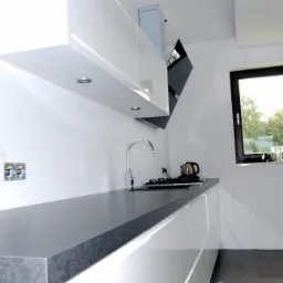 Nowoczesna kuchnia z białymi, lakierowanymi szafkami, szarym blatem z delikatnym wzorem i widokiem na zieleń za oknem, minimalistyczny design.