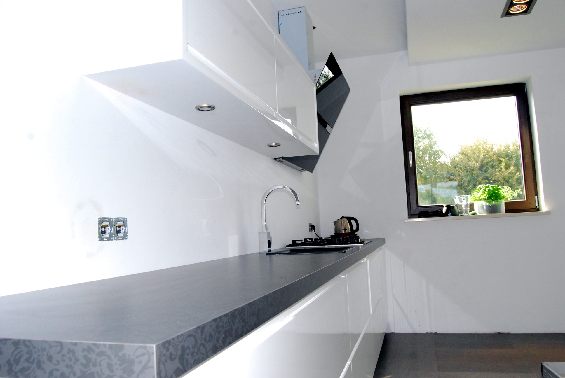 Nowoczesna kuchnia z białymi, lakierowanymi szafkami, szarym blatem z delikatnym wzorem i widokiem na zieleń za oknem, minimalistyczny design.