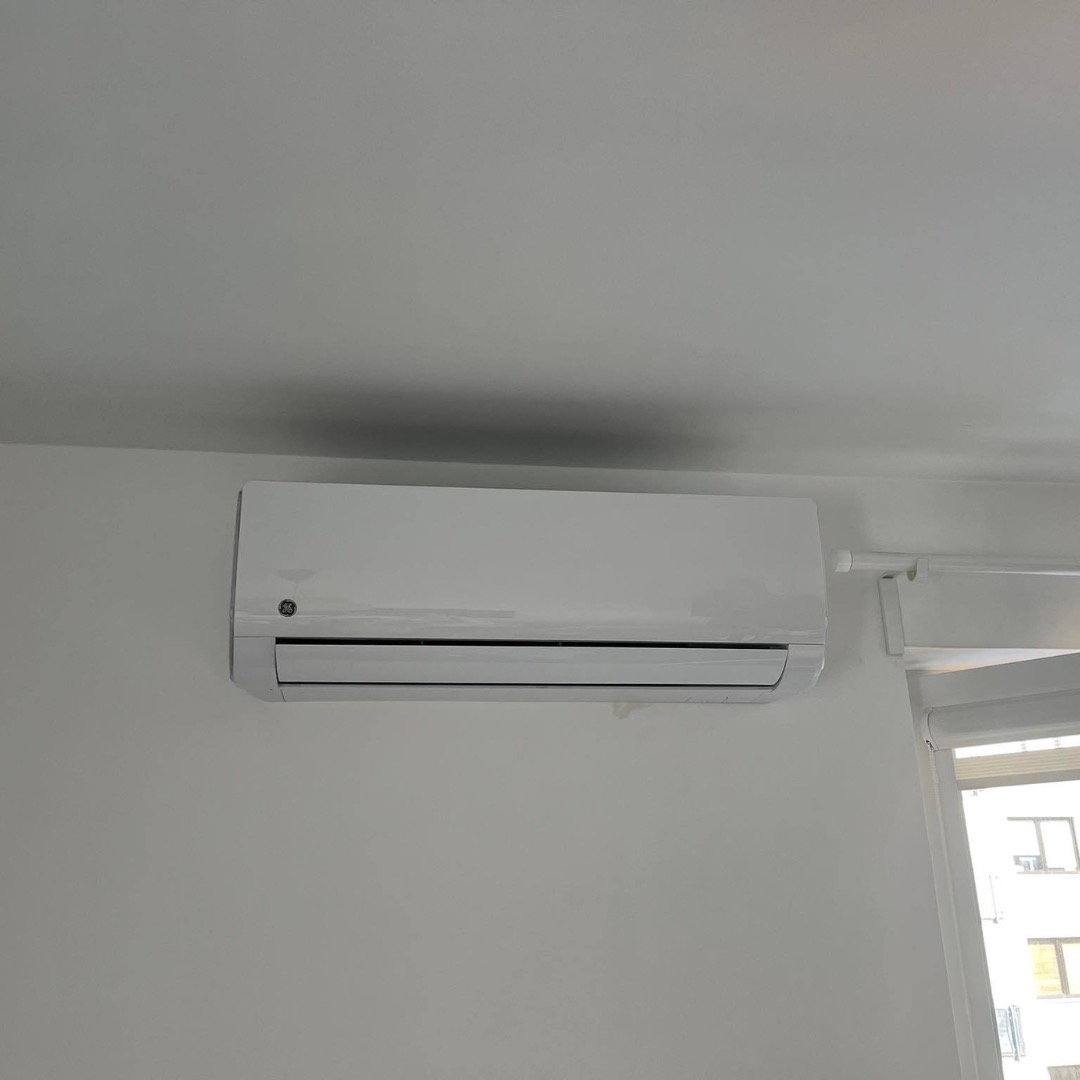 Biały klimatyzator marki GE zamontowany na ścianie w jasnym pokoju z widocznym oknem.