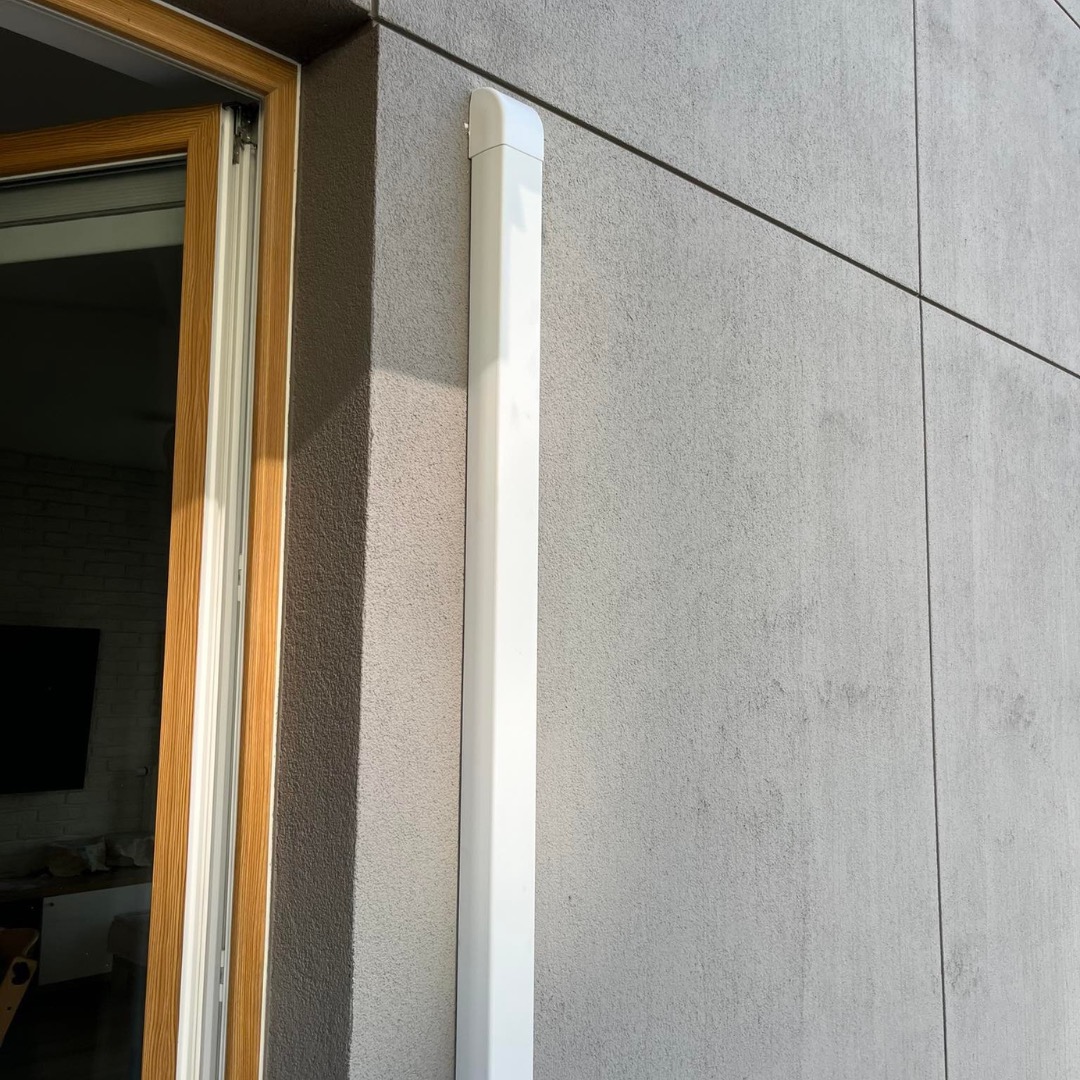 Biała, pionowa maskownica instalacji klimatyzacyjnej na szarej elewacji budynku, widoczna obok okna z drewnianą ramą.