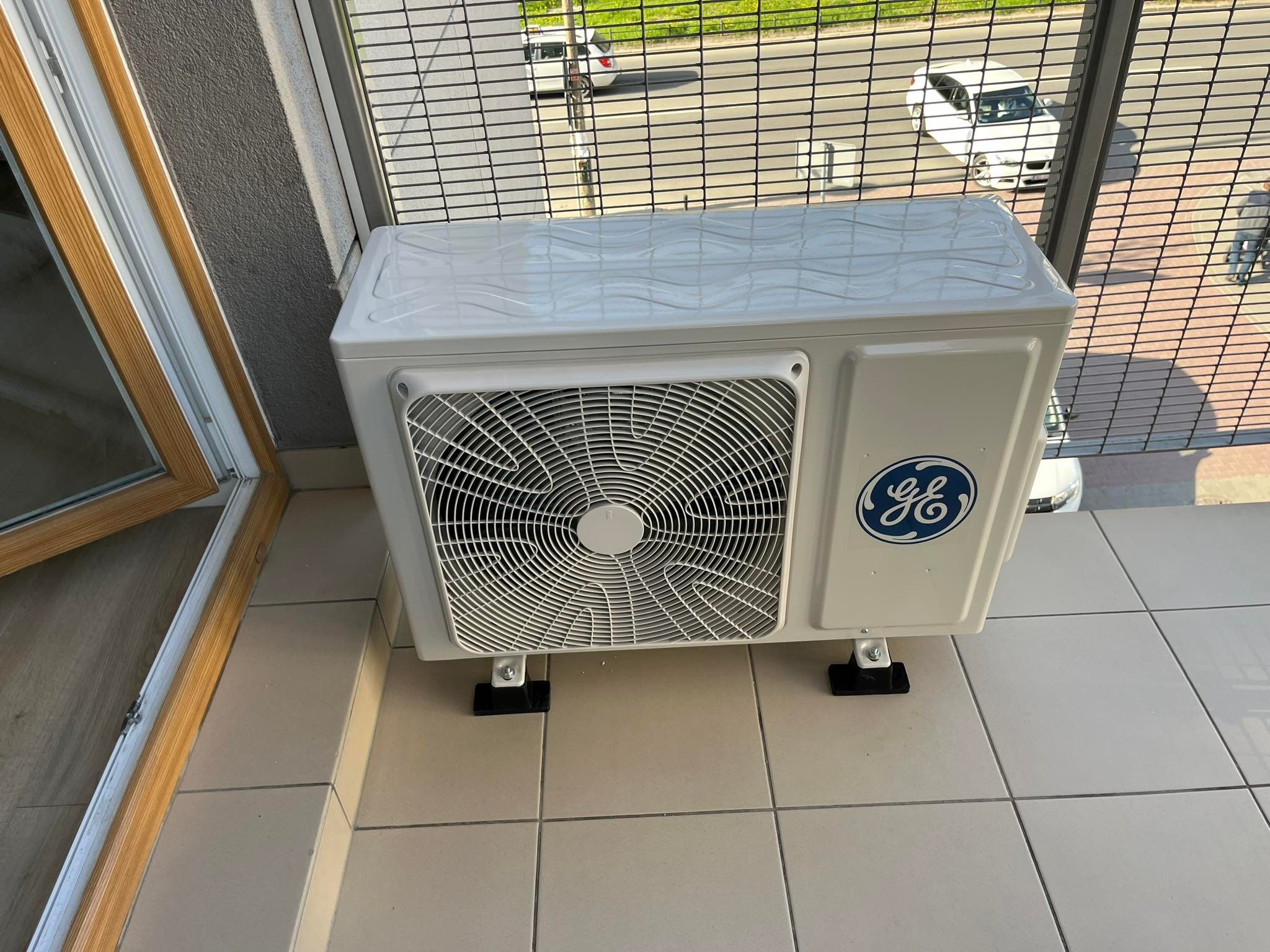 Biała jednostka zewnętrzna klimatyzacji marki GE, zamontowana na balkonie z widokiem na ulicę, widoczna kratka wentylacyjna i logo producenta.