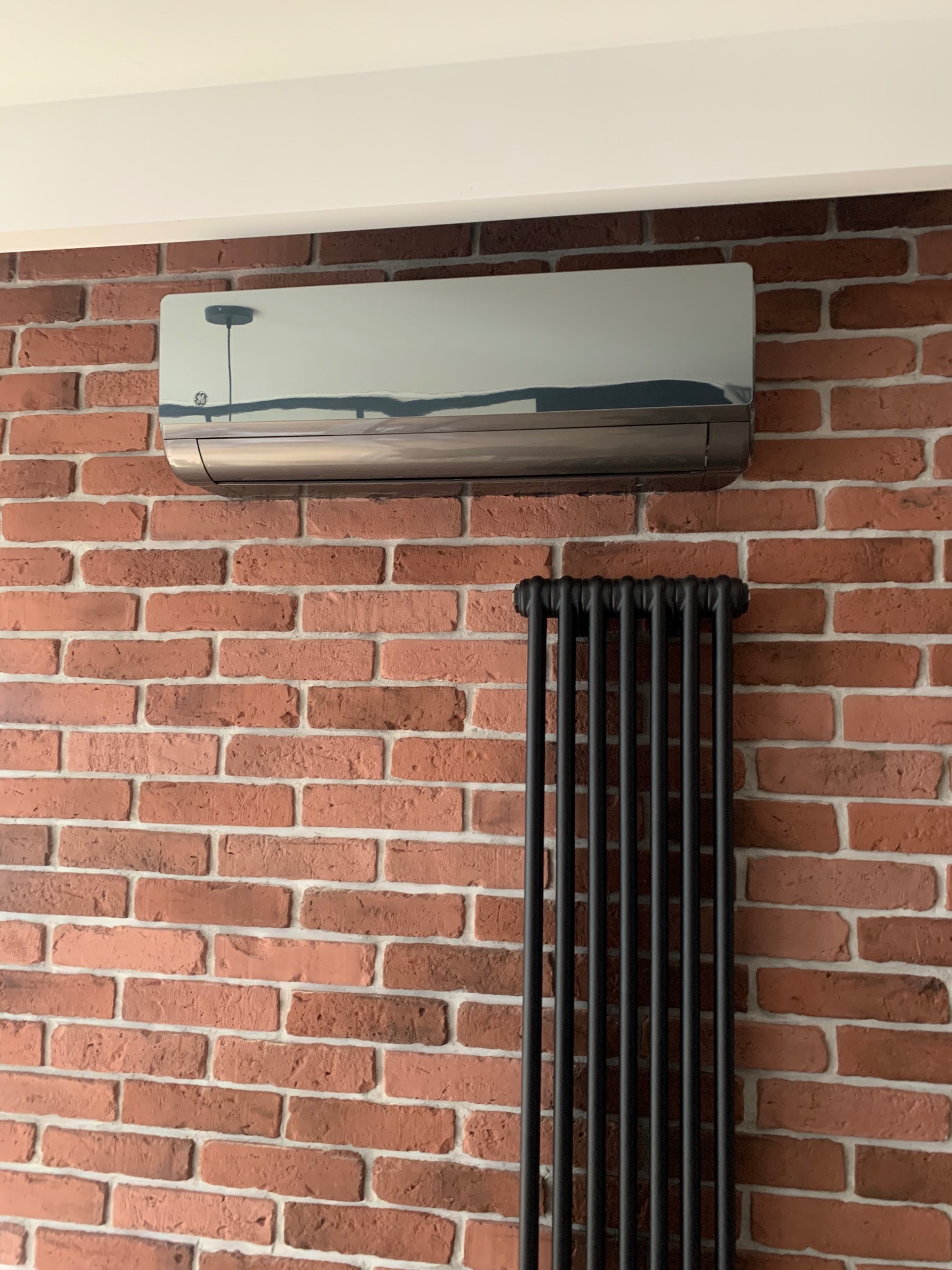 Nowoczesny klimatyzator srebrny zamontowany na ścianie z czerwonej cegły, obok grzejnik żeberkowy w kolorze czarnym.