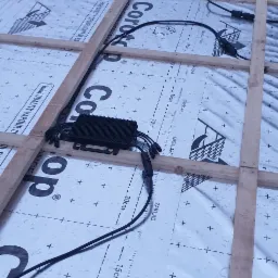 Montaż instalacji fotowoltaicznej na dachu: widoczne czarne skrzynki z przewodami, ułożone na folii paroizolacyjnej z drewnianą konstrukcją nośną.