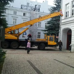 Żółty dźwig samochodowy z napisem 'SPRZĘTBUD 25 T' na tle białego, trzypiętrowego budynku z rusztowaniem i pracującymi na nim osobami. Osoby stoją również na platformie dźwigu.