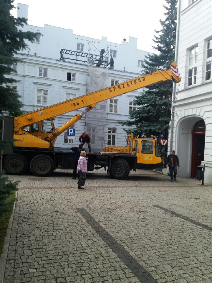Żółty dźwig samochodowy z napisem 'SPRZĘTBUD 25 T' na tle białego, trzypiętrowego budynku z rusztowaniem i pracującymi na nim osobami. Osoby stoją również na platformie dźwigu.