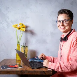 Uśmiechnięta kobieta w okularach i słuchawkach, siedząca przy drewnianym biurku z laptopem, drinkiem i wazonem z żółtymi kwiatami na tle szarej ściany.