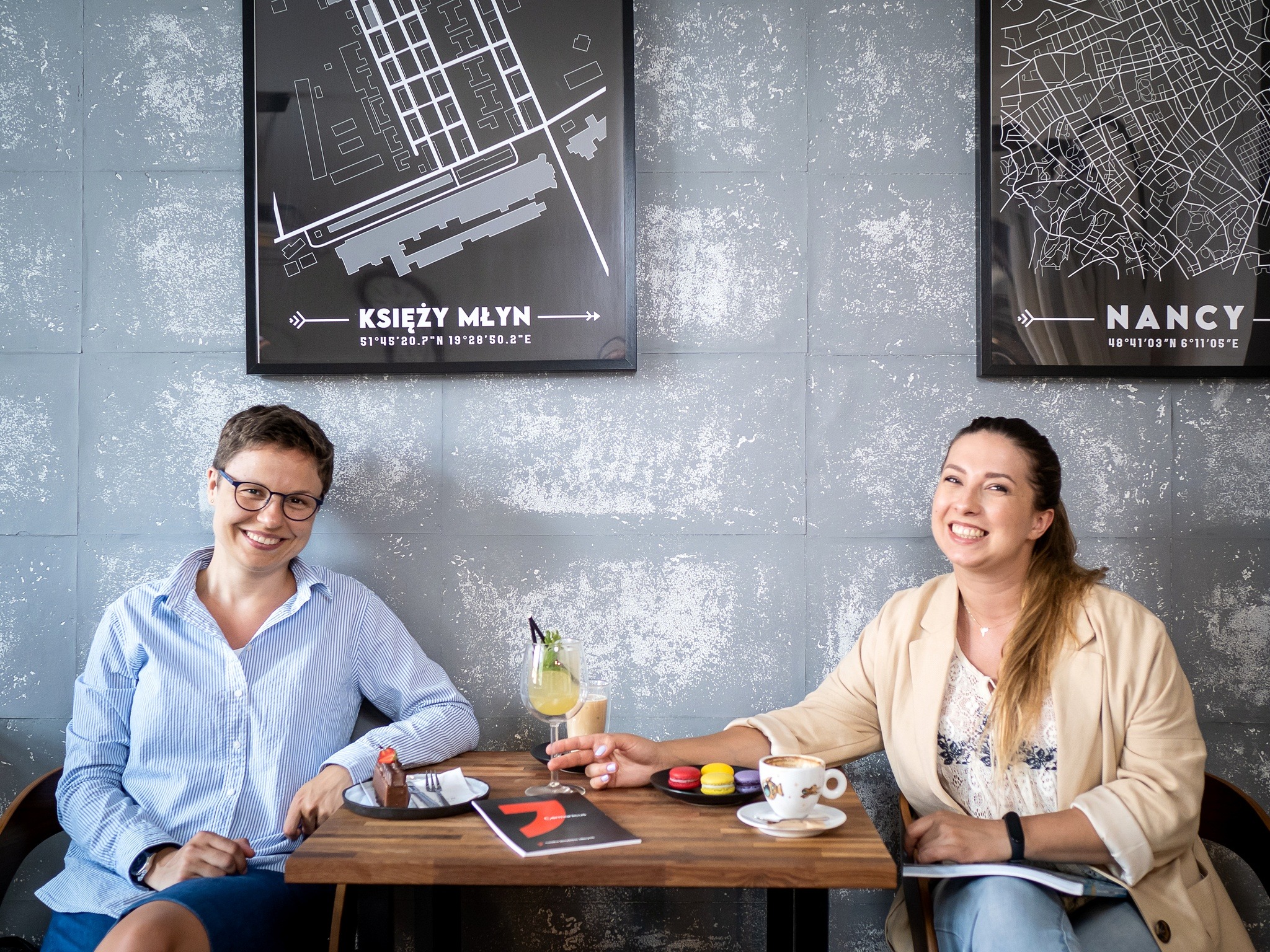 Dwie uśmiechnięte kobiety siedzą przy drewnianym stole w kawiarni, na którym znajdują się desery, napoje i książka z czerwoną okładką; na ścianie wiszą czarno-białe plakaty z mapami.