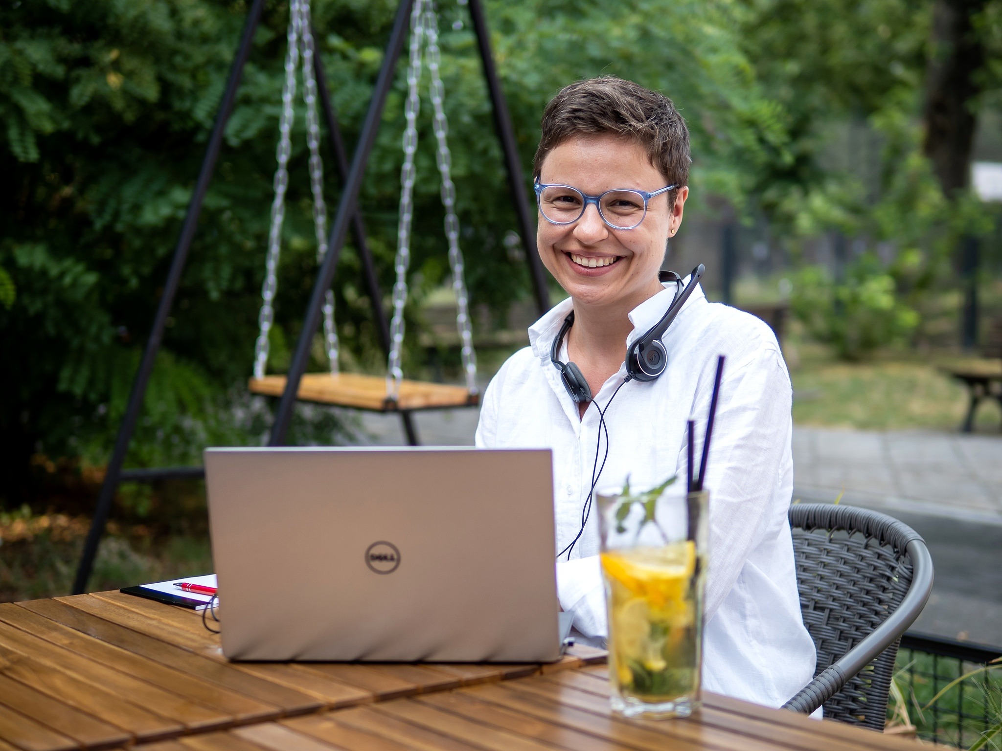 Uśmiechnięta kobieta w okularach i słuchawkach siedzi przy drewnianym stole na zewnątrz z laptopem Dell i szklanką lemoniady, w tle huśtawka łańcuchowa i zieleń.