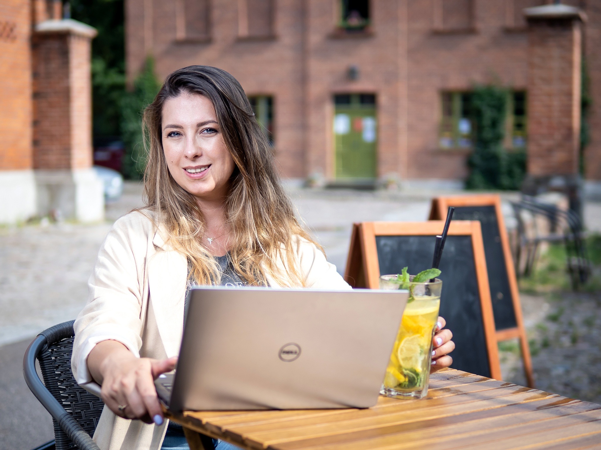Kobieta z laptopem Dell i lemoniadą na drewnianym stole w Łodzi, z ceglanym budynkiem w tle.