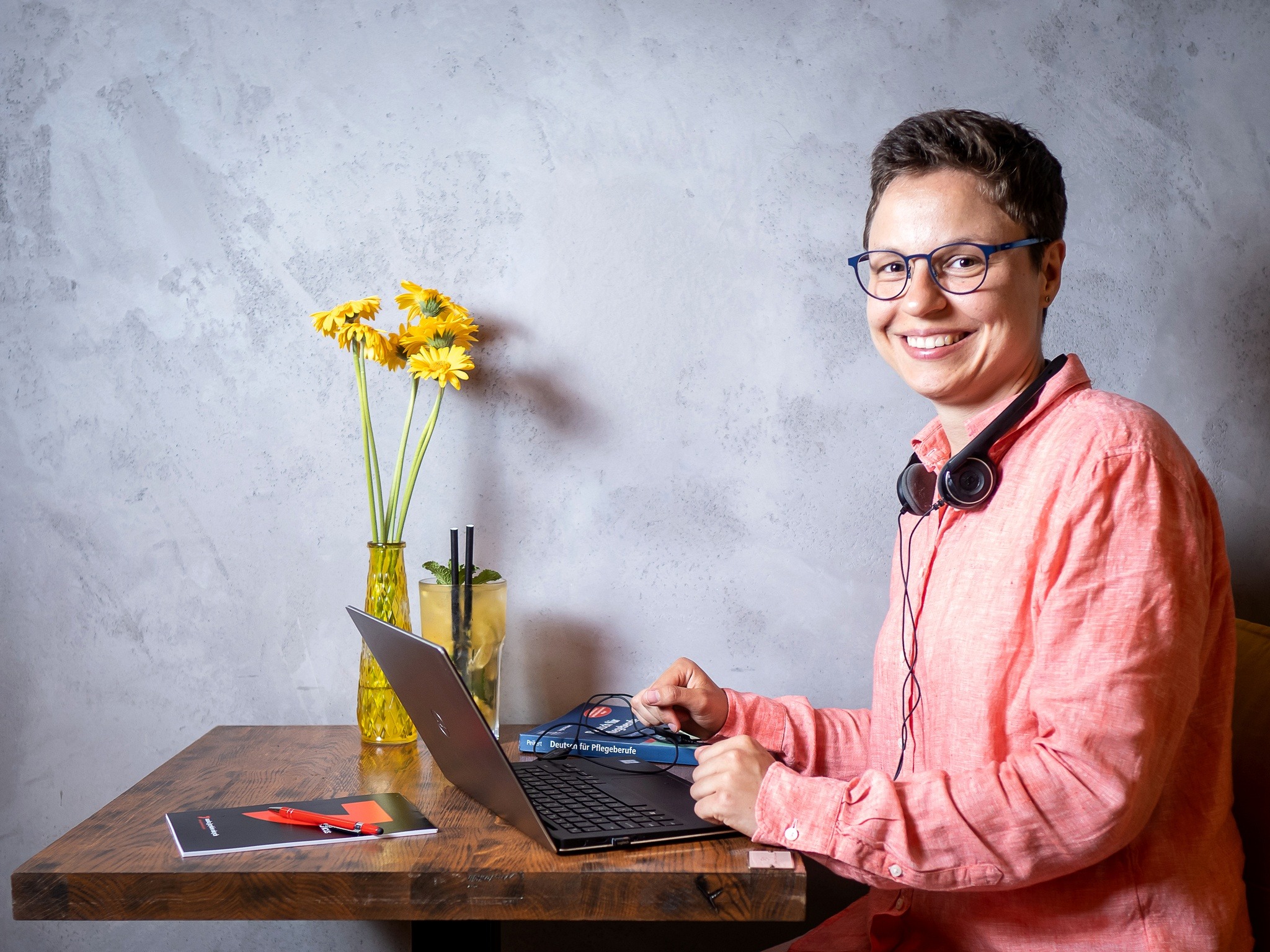 Uśmiechnięta kobieta w okularach i słuchawkach, siedząca przy drewnianym biurku z laptopem, drinkiem i wazonem z żółtymi kwiatami na tle szarej ściany.