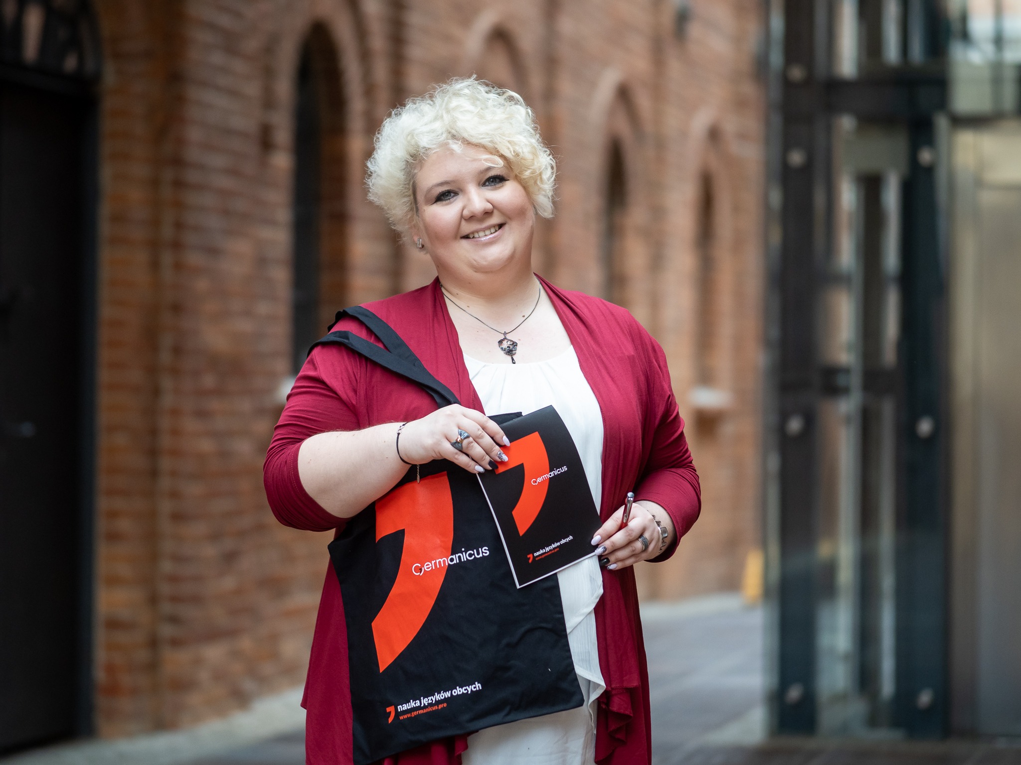 Uśmiechnięta kobieta z blond włosami trzyma torbę i folder z logo szkoły językowej Germanicus na tle ceglanej ściany.