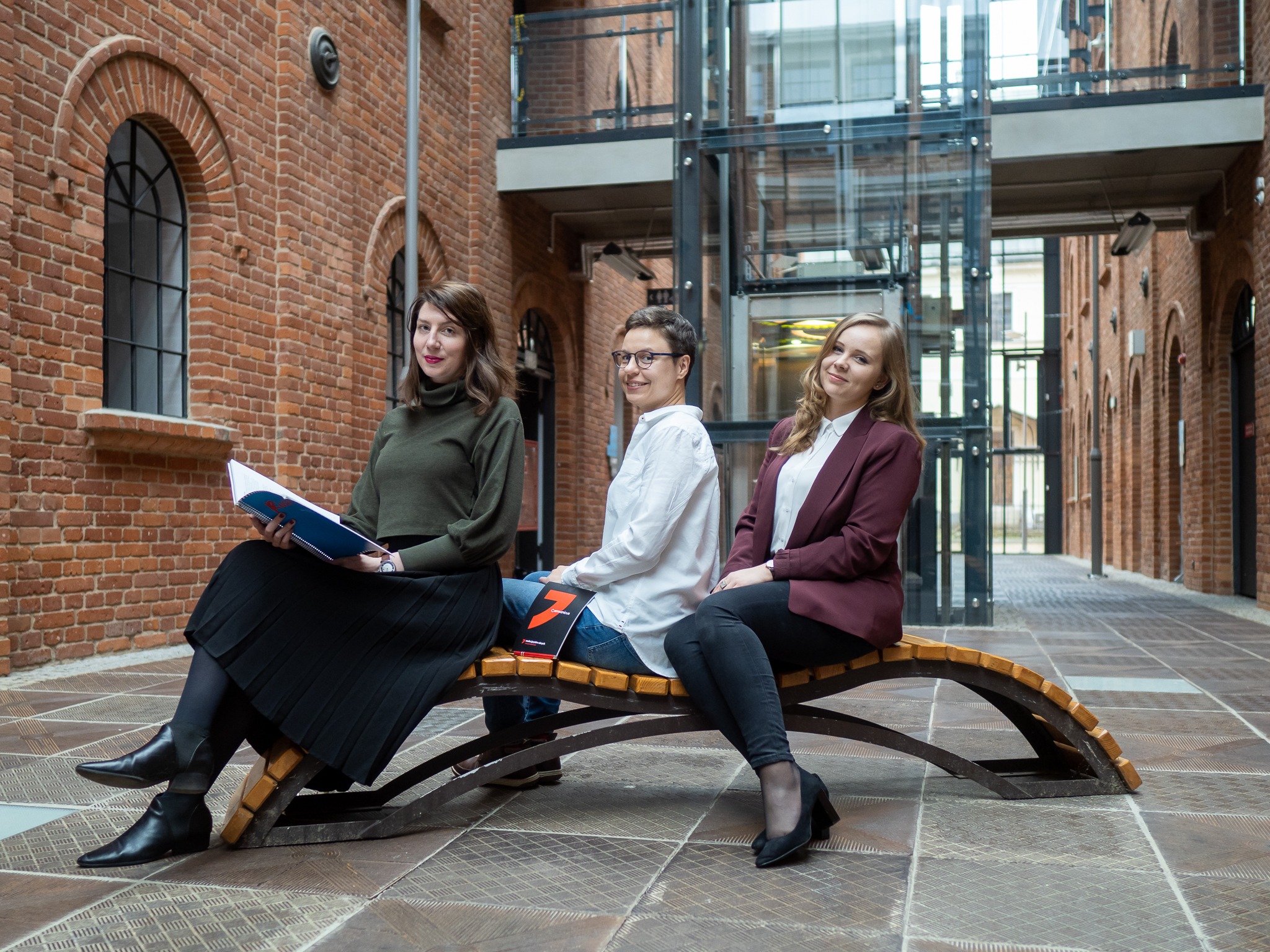 Trzy kobiety siedzą na ławce w przestronnym, ceglanym wnętrzu. Jedna trzyma otwarty notatnik, druga ma okulary i patrzy w obiektyw, trzecia jest ubrana w bordową marynarkę. W tle widoczna...