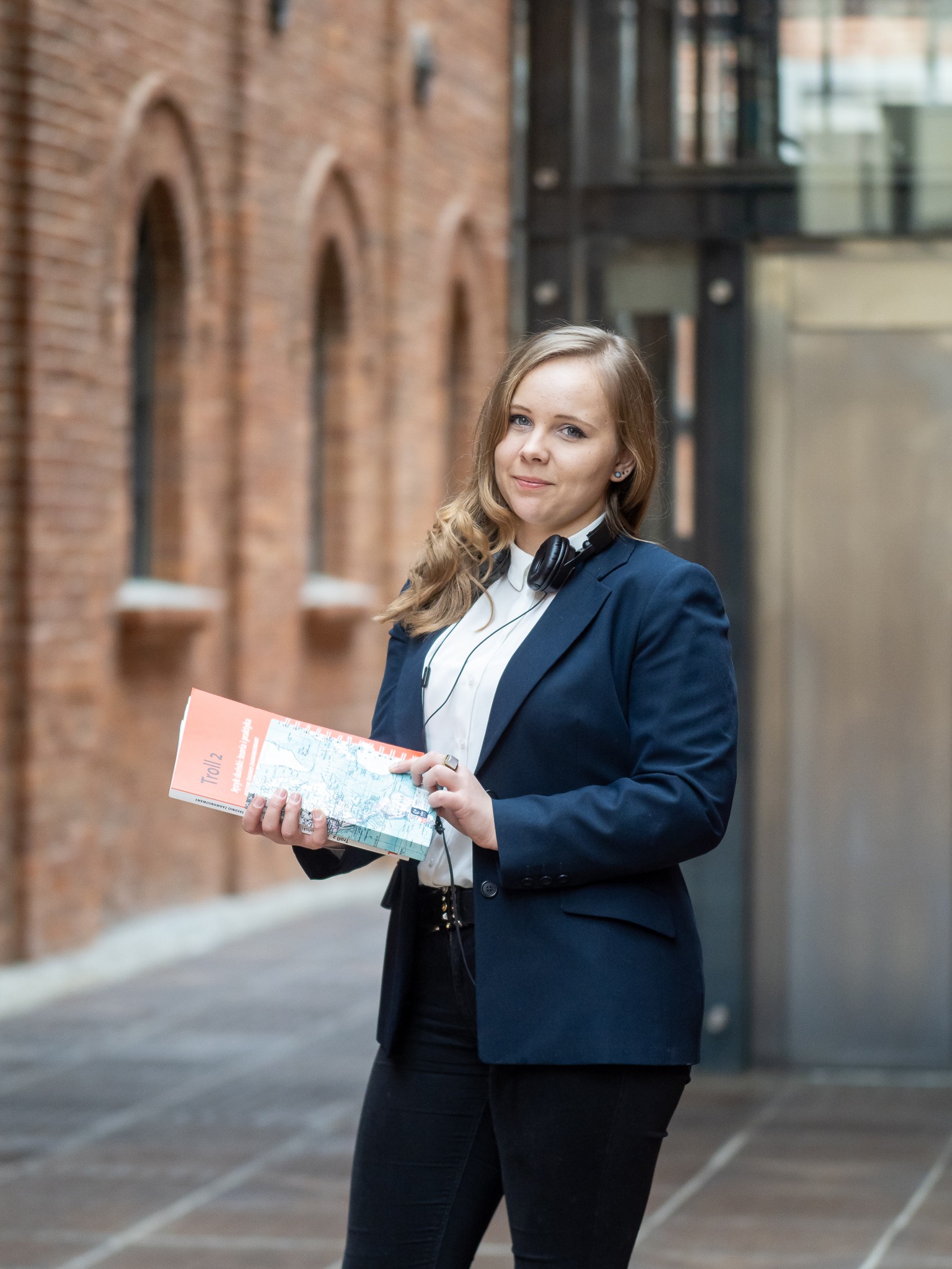 Młoda kobieta w granatowej marynarce i słuchawkach na szyi trzyma w dłoniach podręcznik do języka duńskiego 'Troll 2' na tle ceglanej ściany.