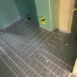 Montaż czerwonych kabli grzewczych na siatce w szarej wylewce podłogowej w surowej łazience z zielonymi płytami kartonowo-gipsowymi na ścianach i wystającymi rurami.