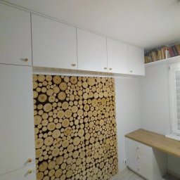 Max furniture - Białe szafki wiszące z drewnianymi gałkami nad ścianą wyłożoną okrągłymi plastrami drewna, obok biurko z drewnianym blatem i szufladami, półka z książkami pod sufitem.