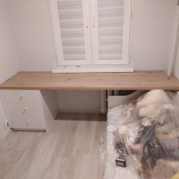 Max furniture - Biurko z blatem drewnianym i białymi szufladami pod oknem, z widocznym fragmentem łóżka piętrowego z zabezpieczeniem i zabawkami przykrytymi folią ochronną.