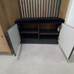 Max furniture - Wnęka z siedziskiem i szafką z otwartymi drzwiczkami w nowoczesnym przedpokoju. Siedzisko z czarną tapicerką, ściana za siedziskiem z pionowych drewnianych listew. Szafka z szarymi drzwiczkami...