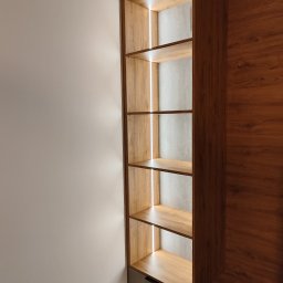 Max furniture - Wysoka, drewniana szafa wnękowa z oświetleniem LED wewnątrz półek, z szufladami na dole, dopasowana kolorystycznie do panelu ściennego po prawej stronie.