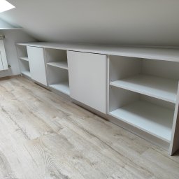 Max furniture - Biała zabudowa ze szafkami i półkami, dopasowana do skosu poddasza, z przesuwnymi drzwiami, na podłodze panele w odcieniach beżu i szarości, obok grzejnik.