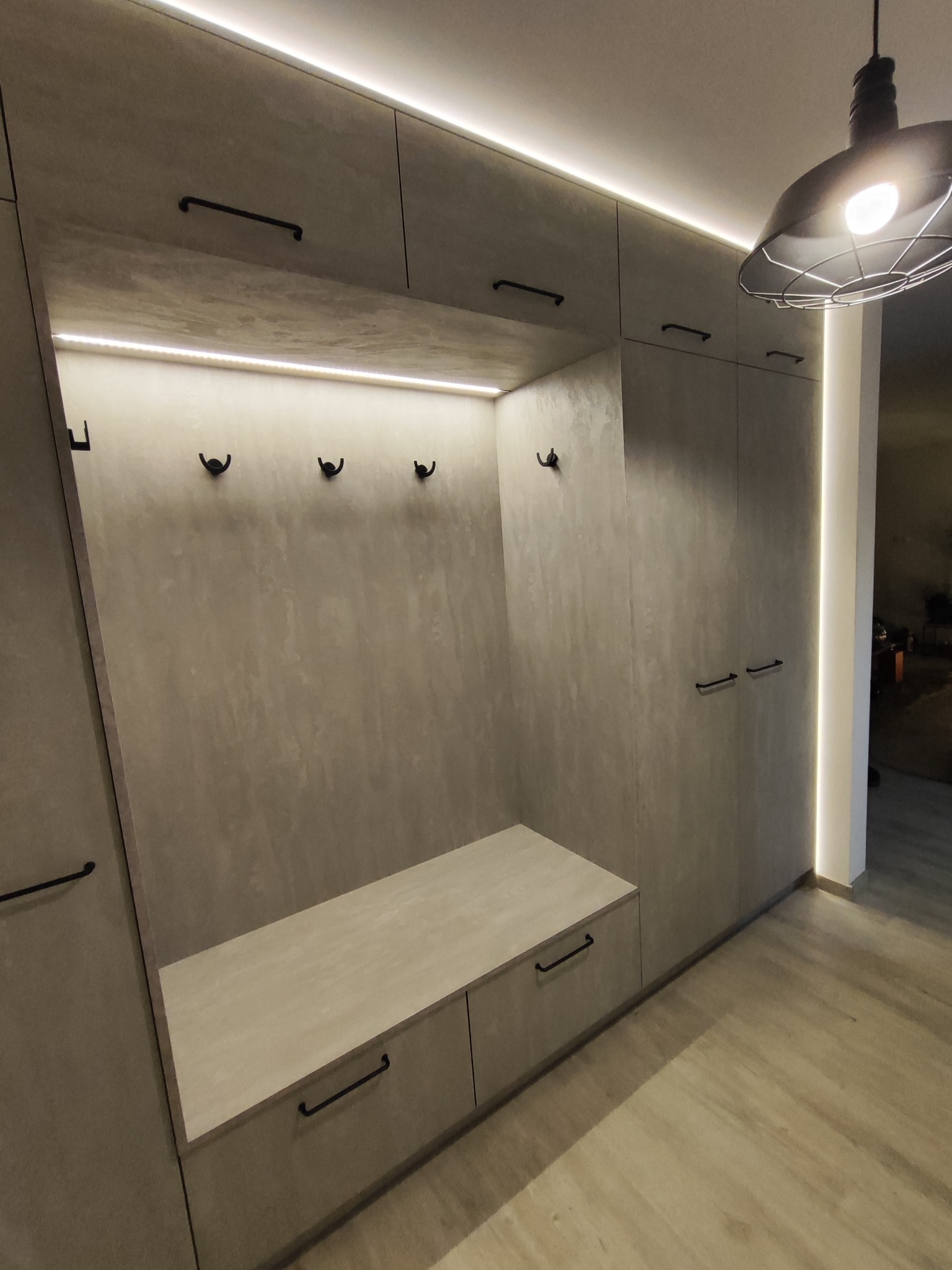 Nowoczesna zabudowa przedpokoju z szarego betonu architektonicznego, z siedziskiem, wieszakami i oświetleniem LED, uzupełniona o metalową lampę wiszącą.