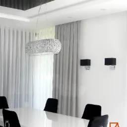 Nowoczesna jadalnia z białym stołem, czarnymi krzesłami z srebrnymi pierścieniami, kryształowym żyrandolem i szarymi zasłonami w tle.