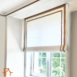 Elegancka, biała roleta rzymska z brązowym obramowaniem, zamontowana w oknie z widokiem na zieleń, częściowo zwinięta, z widocznym logo firmy w lewym dolnym rogu.