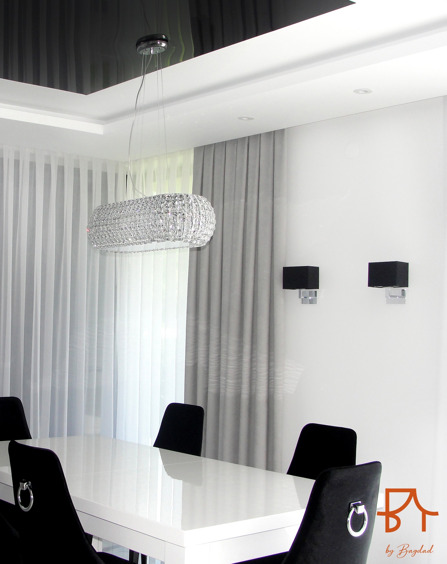 Nowoczesna jadalnia z białym stołem, czarnymi krzesłami z srebrnymi pierścieniami, kryształowym żyrandolem i szarymi zasłonami w tle.