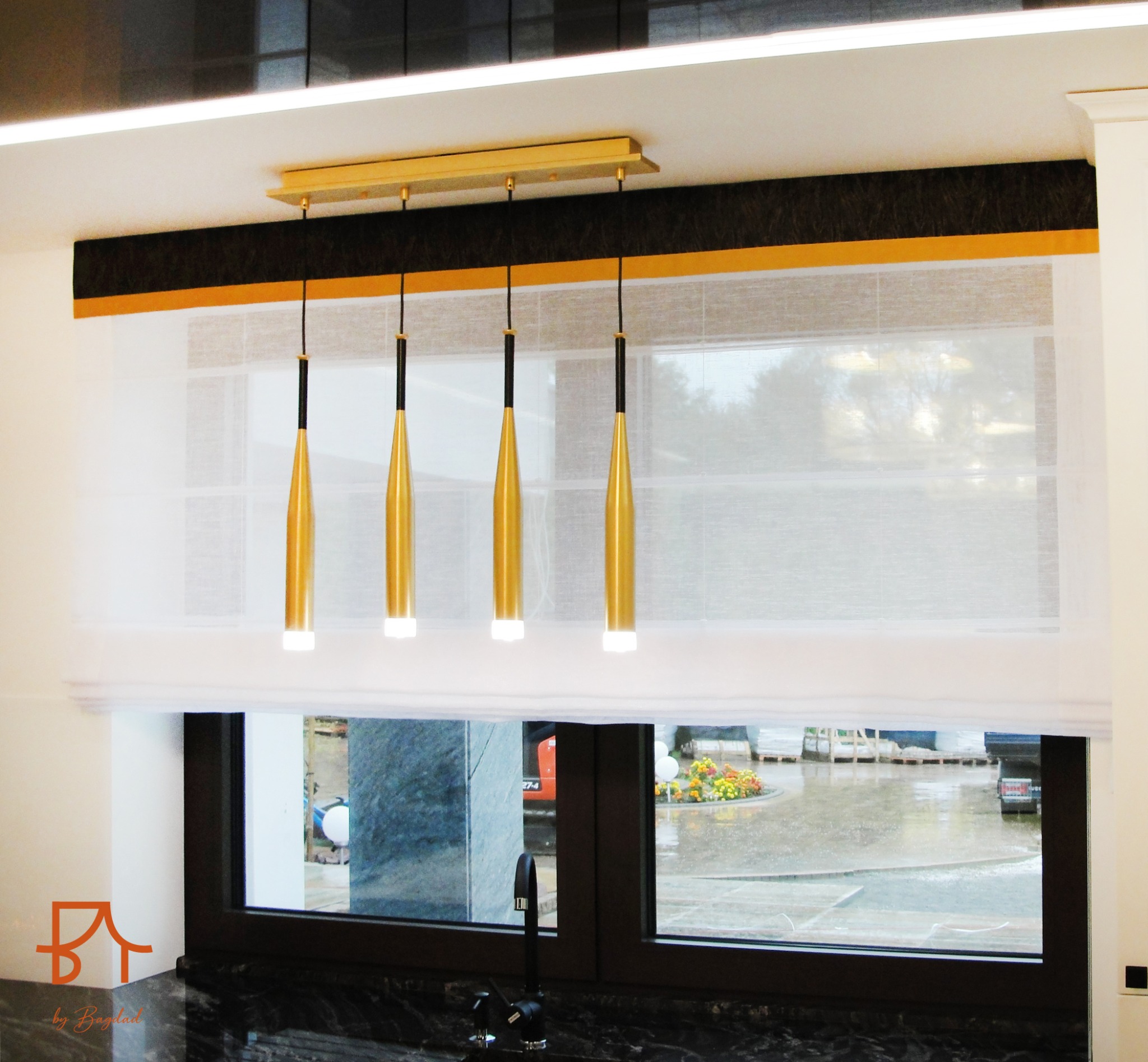 Elegancka, jasna kuchnia z marmurowym blatem i oknem z roletą rzymską, nad którym wiszą trzy złote lampy w kształcie stożków.