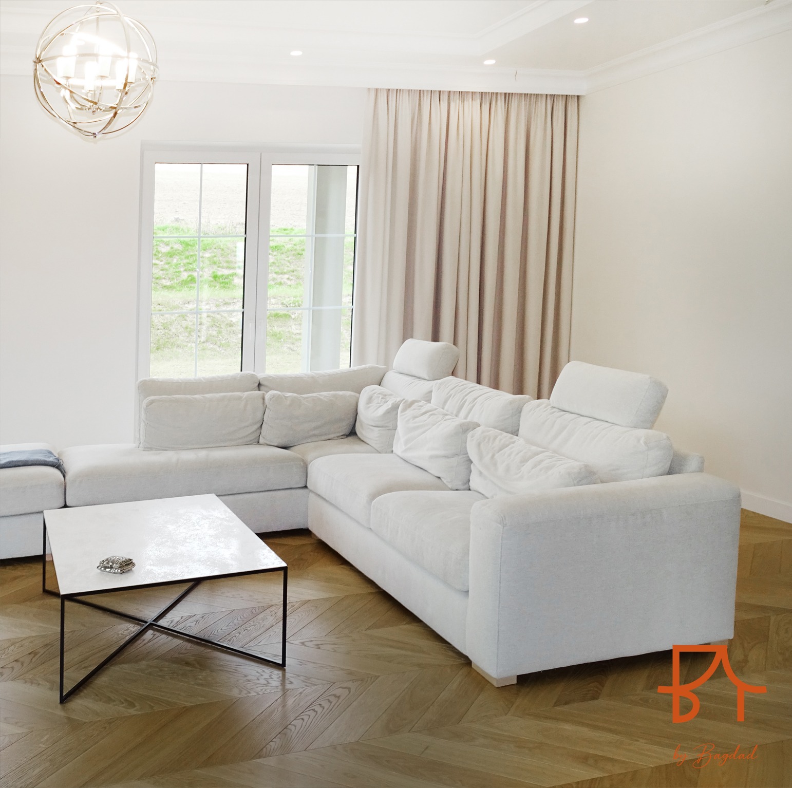 Jasny salon z narożną sofą, stolikiem kawowym z metalową podstawą, parkietem w jodełkę i zasłonami w oknie, całość utrzymana w neutralnej kolorystyce z akcentami bieli.