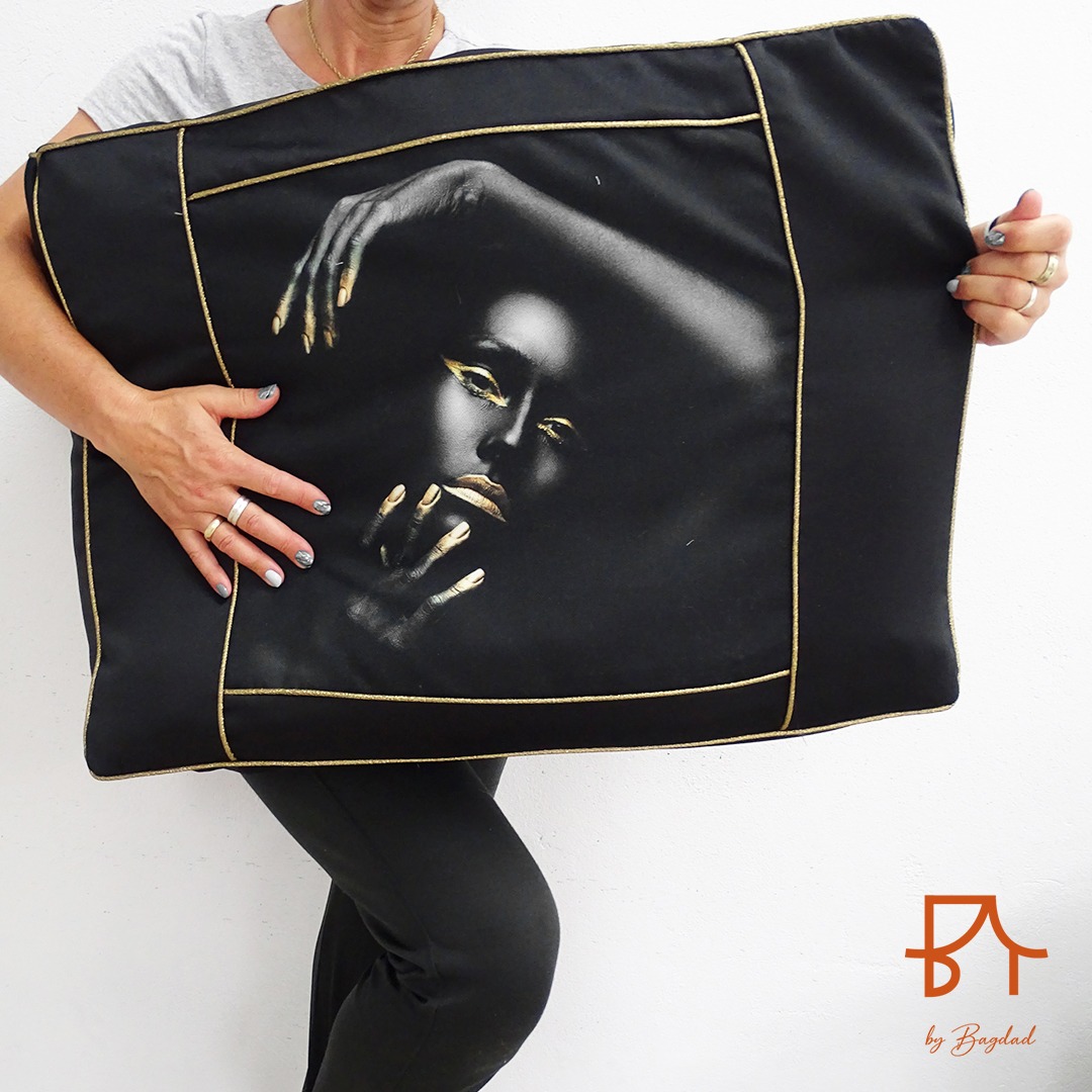 Osoba trzyma czarną poduszkę z nadrukiem portretu kobiety z elementami w kolorze złotym, obramowanie poduszki w kolorze złotym.