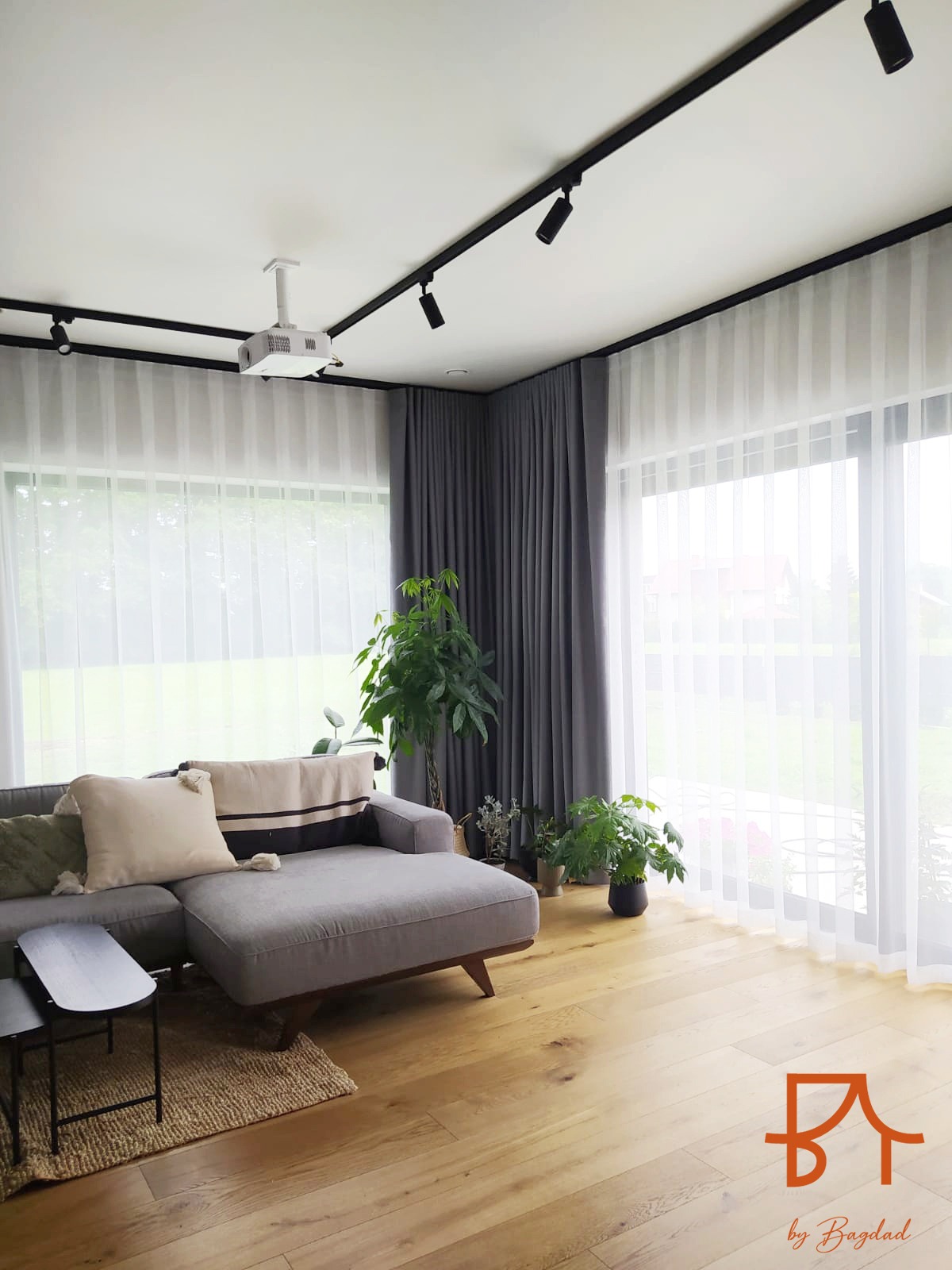 Jasne wnętrze salonu w Krakowie z drewnianą podłogą, szarą sofą i dużymi oknami zasłoniętymi białymi firanami i szarymi zasłonami, oświetlone reflektorami na szynoprzewodzie i z projektorem...