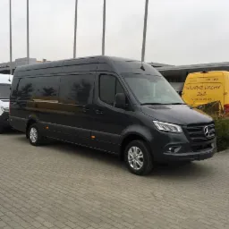 Czarny, długi bus Mercedes Sprinter na brukowanym placu, z żółtym busem w tle i słupami oświetleniowymi. Widoczne logo Mercedesa na masce.