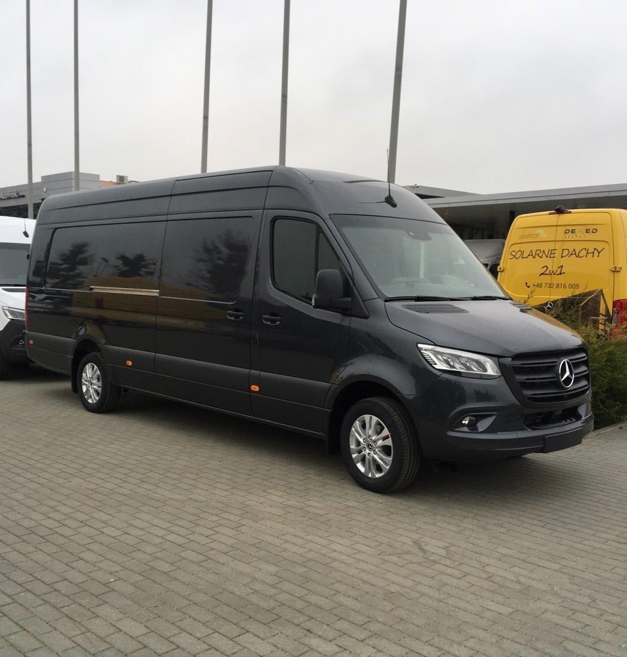 Czarny, długi bus Mercedes Sprinter na brukowanym placu, z żółtym busem w tle i słupami oświetleniowymi. Widoczne logo Mercedesa na masce.