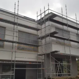 Ocieplanie i wykańczanie elewacji dwukondygnacyjnego budynku z płaskim dachem, widoczne rusztowania i pracownicy w odblaskowych kamizelkach.