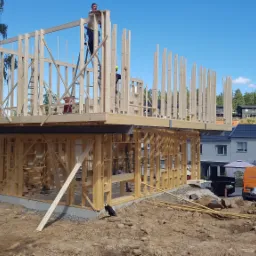 Wznoszenie dwukondygnacyjnej konstrukcji szkieletowej domu z widocznymi pracownikami na rusztowaniu i wewnątrz budynku, z mini-wywrotką i pomarańczowym vanem w tle.