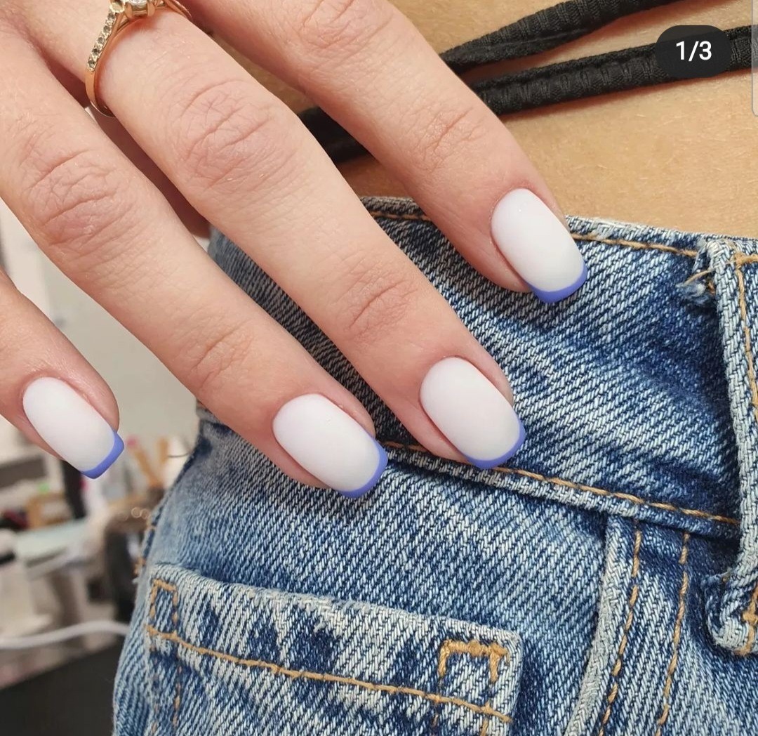 Dłoń z białymi, matowymi paznokciami z fioletową obwódką, na tle dżinsowej kieszeni.